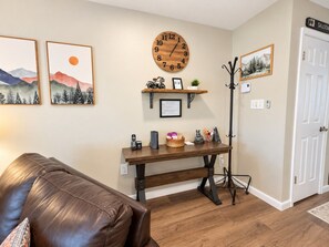 Una sala de estar con un sofá de cuero marrón, una mesa de centro de madera con varios objetos, un reloj de pared y dos cuadros enmarcados.