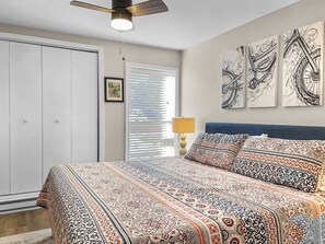 A bedroom with a bed, a ceiling fan, a window with blinds, a lamp, and artwork on the wall.