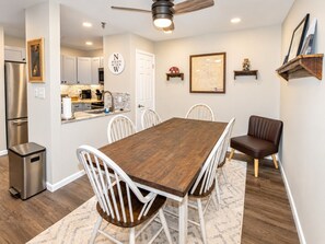A dining area with a wooden table and white chairs, a kitchen in the background, and a ceiling fan.