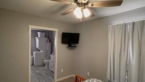 A bedroom with a ceiling fan, a bed with a patterned bedspread, a television on the wall, and a bathroom visible through an open door. - Little Country House a Quiet Countryside Getaway! (Newton)