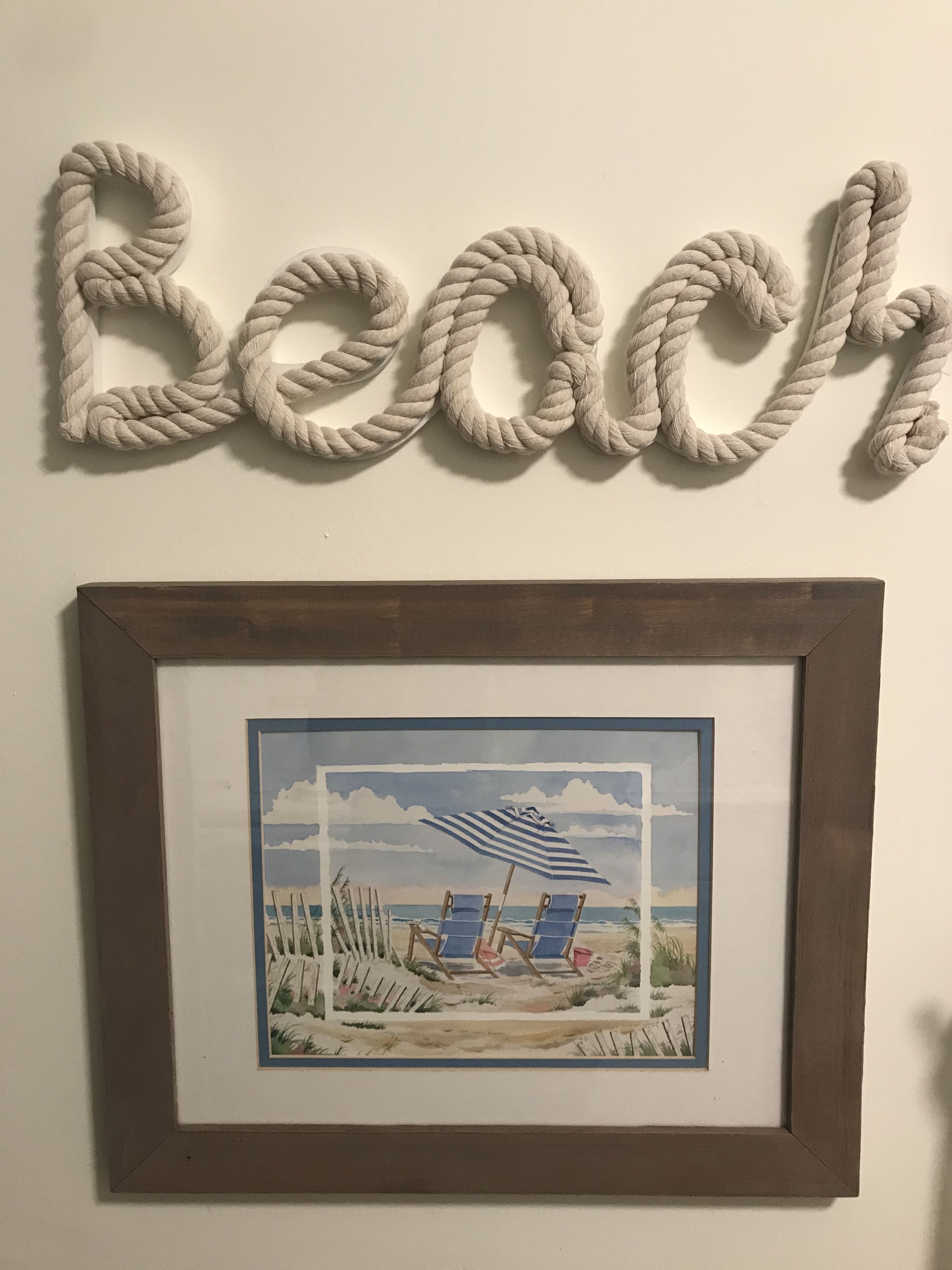 A framed beach scene with two chairs and an umbrella, mounted on a wall above the word "Beach" spelled out in rope letters.