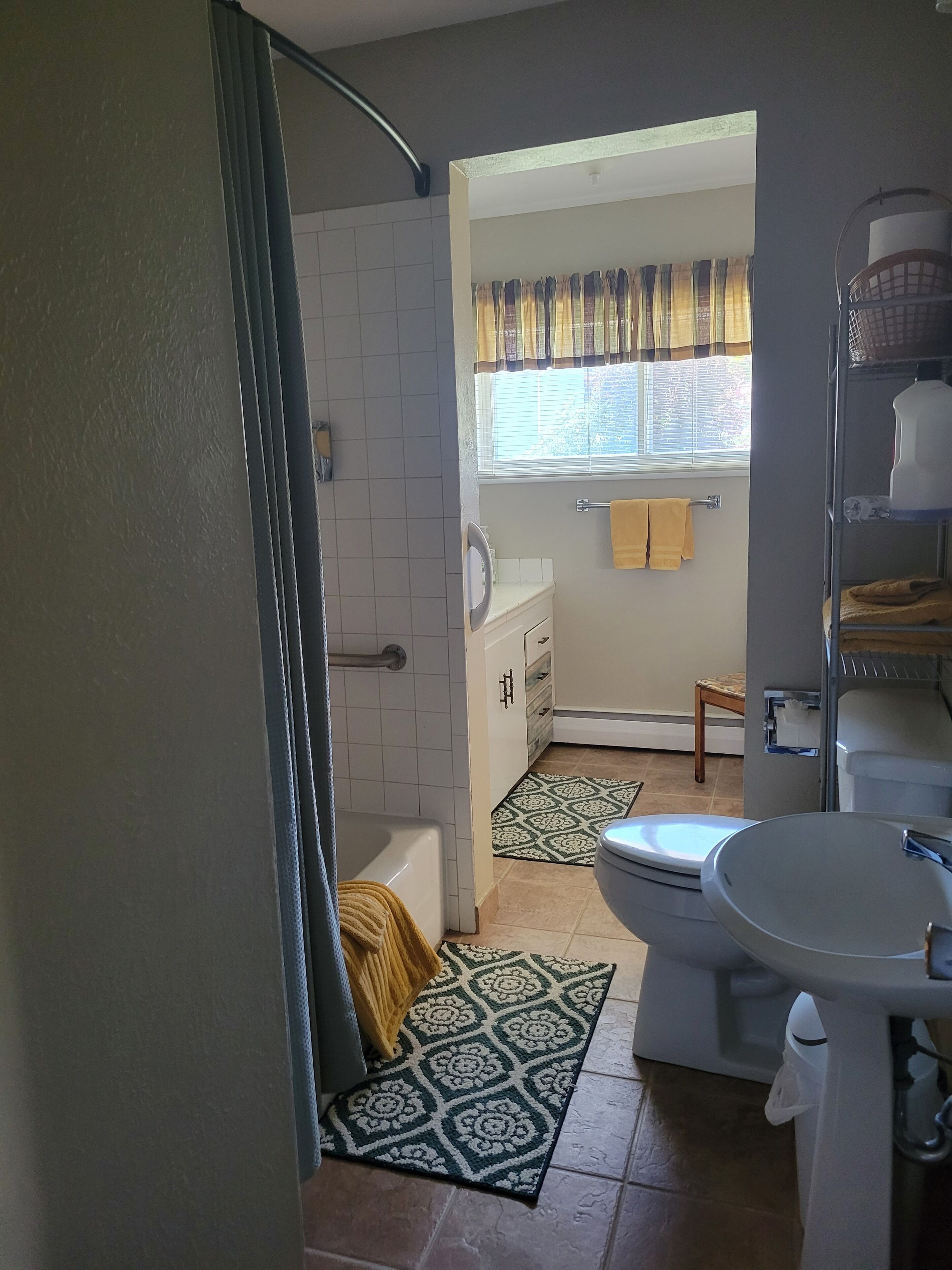Et badeværelse med badekar, toilet og vask. Der er et vindue med gardiner, et håndklædestativ og en gulvtæppe med et mønster.