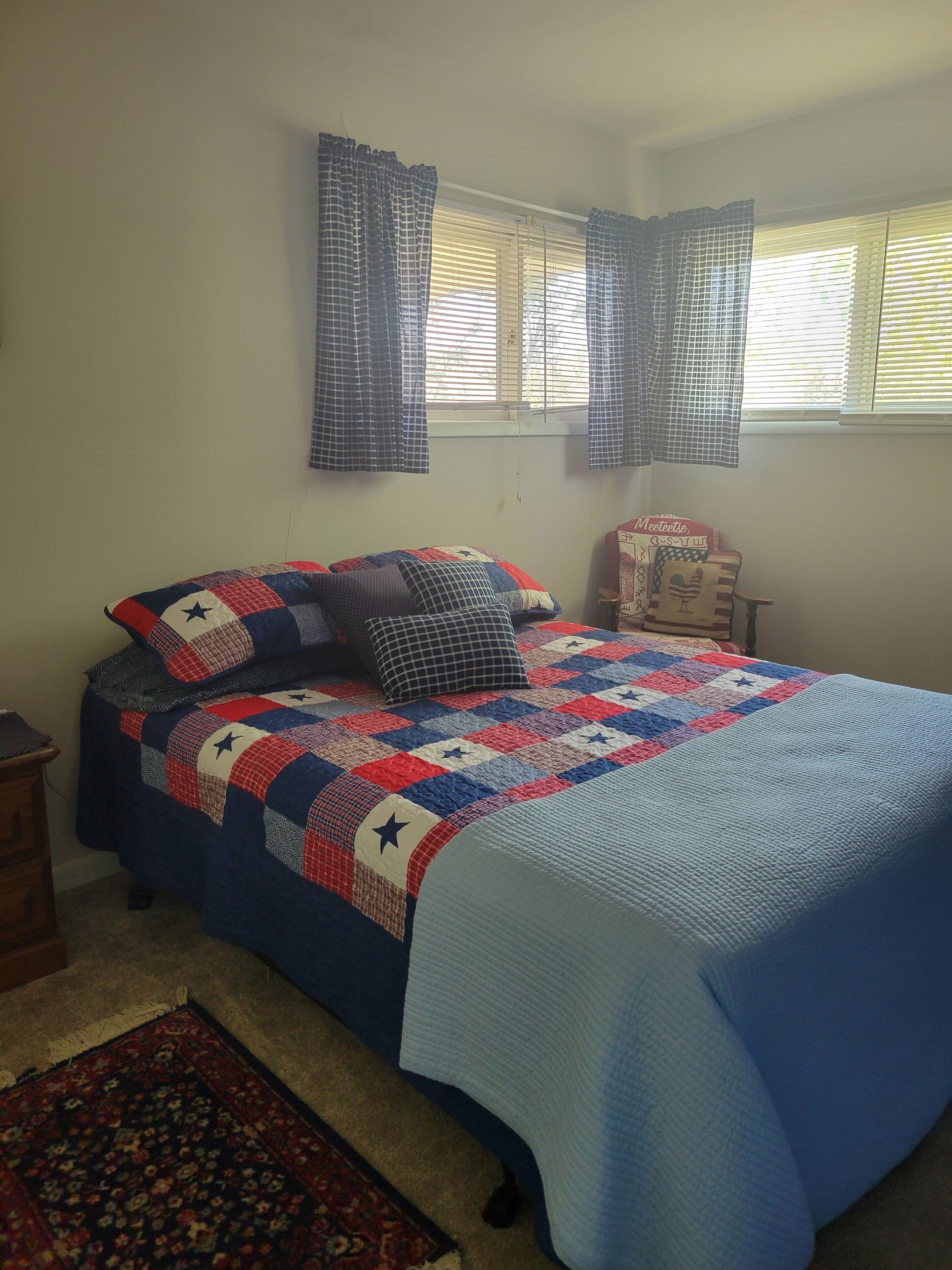 A bedroom with a bed covered in a patchwork quilt, a wooden nightstand, a chair with a cushion, and a window with blinds.