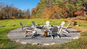 A circular fire pit with Adirondack chairs around it, set on a gravel area with trees in the background. - Beautiful Historic Country Estate – Modern Comfort in Okemo Valley (Cavendish)