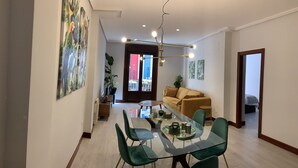 A modern dining area with a glass table, green chairs, and a chandelier. There is a sofa in the background and a door leading to another room.
