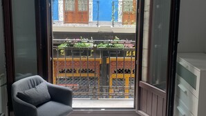 A room with a bed, a chair, a wardrobe, and a balcony with a view of buildings and a blue sky.