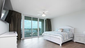 A bedroom with a bed, a nightstand, a TV, a ceiling fan, and a sliding glass door leading to a balcony. - Luxurious Caribe bay-view condo, 4BR/3BA, sleeps 9 (Orange Beach)