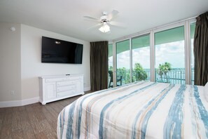 Habitación de hotel con cama, televisor montado en la pared, cómoda blanca y balcón con vista al océano.