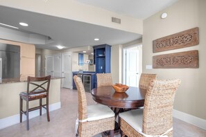 A dining area with a round table and four wicker chairs, a kitchen in the background, and decorative wall art. - Cozy Nautical Condo Overlooking Perdido Bay! Complimentary Boat Slip! (Orange Beach)