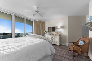 Una habitación de hotel con cama, una silla, cómoda, ventilador de techo y un ventanal grande.