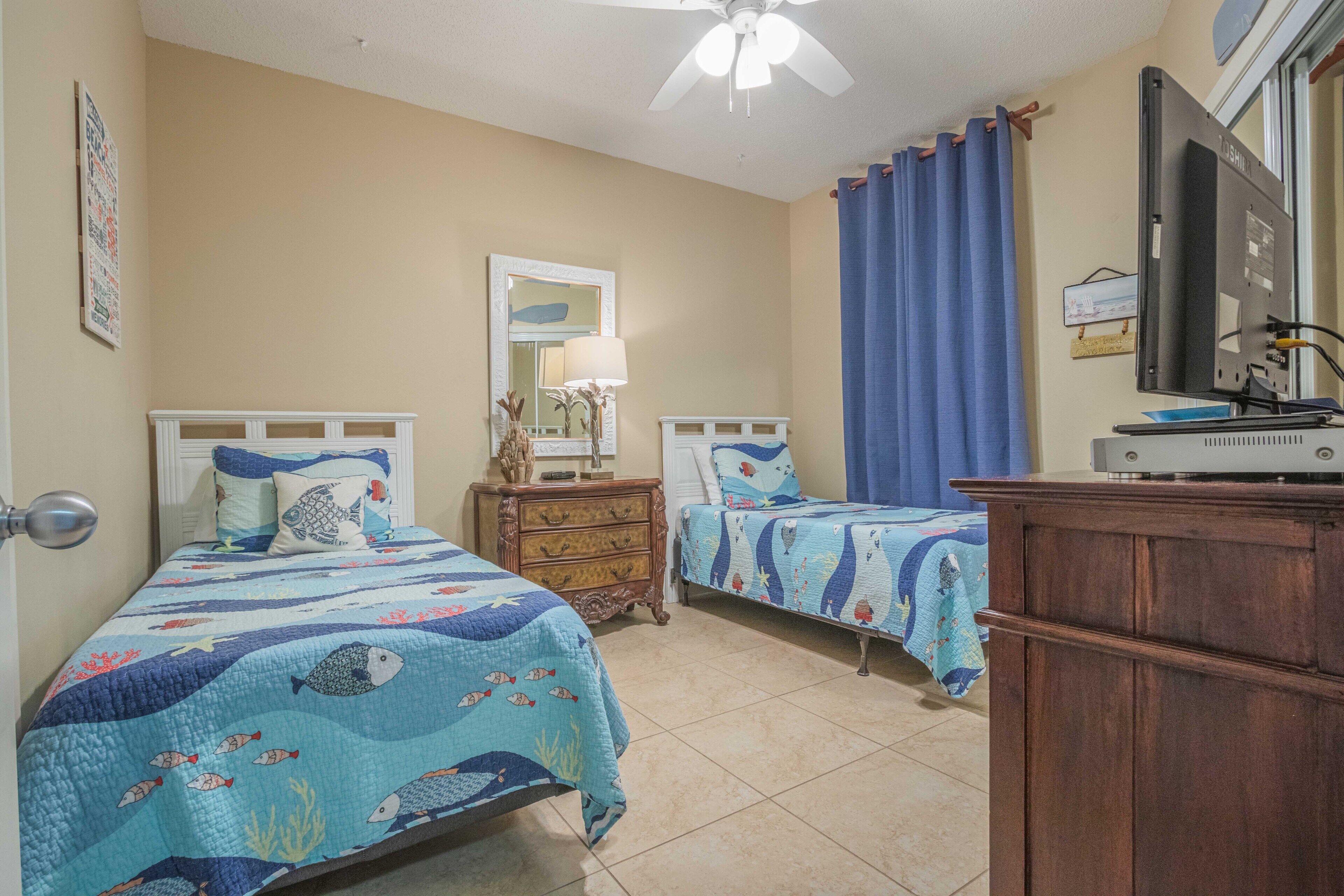 Quarto com duas camas, cômoda, ventilador de teto, televisão e janela com cortinas.