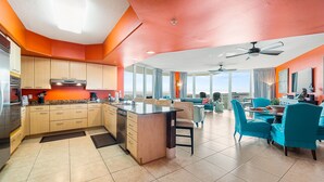 A modern kitchen with a central island, a dining area with blue chairs, and a large window offering a view. - Luxurious Caribe 4BR/3BA Beach View Condo (Orange Beach)