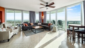 A spacious living room with a sectional sofa, a coffee table, and a dining area with a table and chairs. Large windows offer a view of the outdoors. - Luxurious Caribe 4BR/3BA Beach View Condo (Orange Beach)