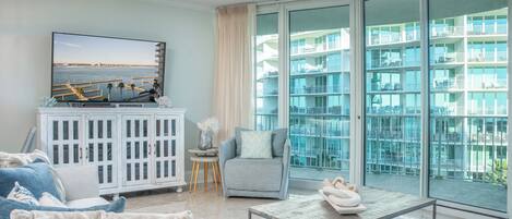 A modern living room with a television, a sofa, a chair, a coffee table, and a view of a balcony with a pool and buildings.