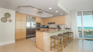 A modern kitchen with a stainless steel refrigerator, wooden cabinets, a central island with bar stools, and a view of the ocean through large windows. - Exquisite Corner Condo Overlooking Outdoor Pools and Perdido Pass! Complimentary Boat Slip! (Orange Beach)