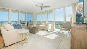 A spacious living room with a large window overlooking the ocean, a ceiling fan, and a television on a wooden cabinet. - Exquisite Corner Condo Overlooking Outdoor Pools and Perdido Pass! Complimentary Boat Slip! (Orange Beach)