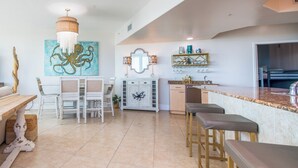 A spacious kitchen with a dining area, featuring a chandelier, a large octopus painting, and a bar with stools. - Exquisite Corner Condo Overlooking Outdoor Pools and Perdido Pass! Complimentary Boat Slip! (Orange Beach)
