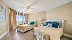 A hotel room with two beds, beige walls, a ceiling fan, a large window with curtains, and a painting on the wall.