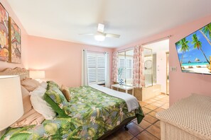 Un dormitorio con cama, mesita de noche, ventana con contraventanas, ventilador de techo y vista a una playa a través de un ventanal.