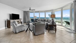 Un salón amplio con un televisor de pantalla plana grande, un sofá y un sillón. La habitación tiene ventanas grandes que ofrecen vistas al océano.