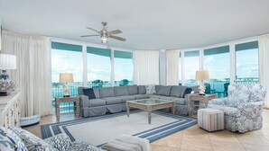 Een ruime woonkamer met een grote grijze hoekbank, een glazen salontafel en een fauteuil met een patroon. De kamer heeft grote ramen met uitzicht op de oceaan en een balkon met een hekwerk.