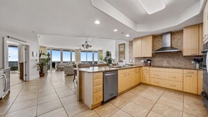 A spacious kitchen with a central island, wooden cabinets, and a dishwasher. There are potted plants and a view of the outdoors through large windows.