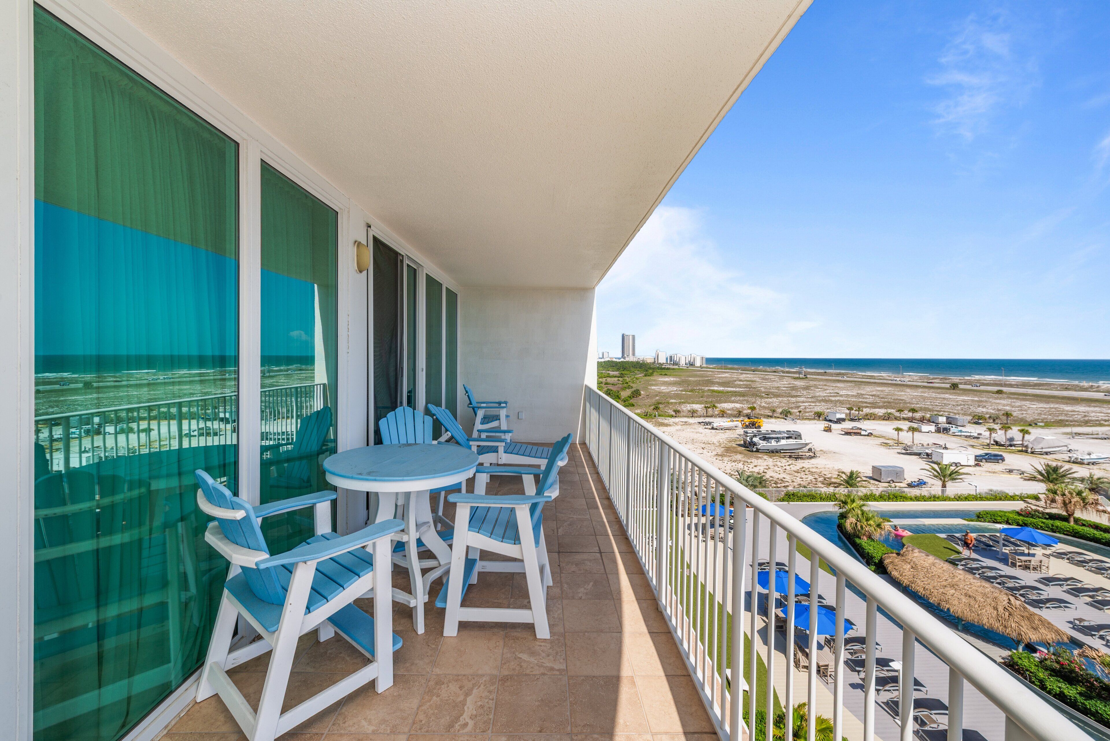 Un balcón con vistas a la playa, una zona de piscina y un comedor con sillas y mesas.