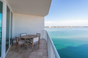 Ein Balkon mit einem runden Holztisch und vier Stühlen, mit Blick auf ein klares blaues Meer und eine entfernte Küste mit Gebäuden.