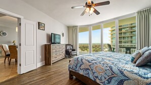 Ein Schlafzimmer mit einem großen Bett, einem Deckenventilator, einem Fernseher, einem Schminktisch, einem Stuhl und einem Balkon mit Blick auf ein Gebäude.