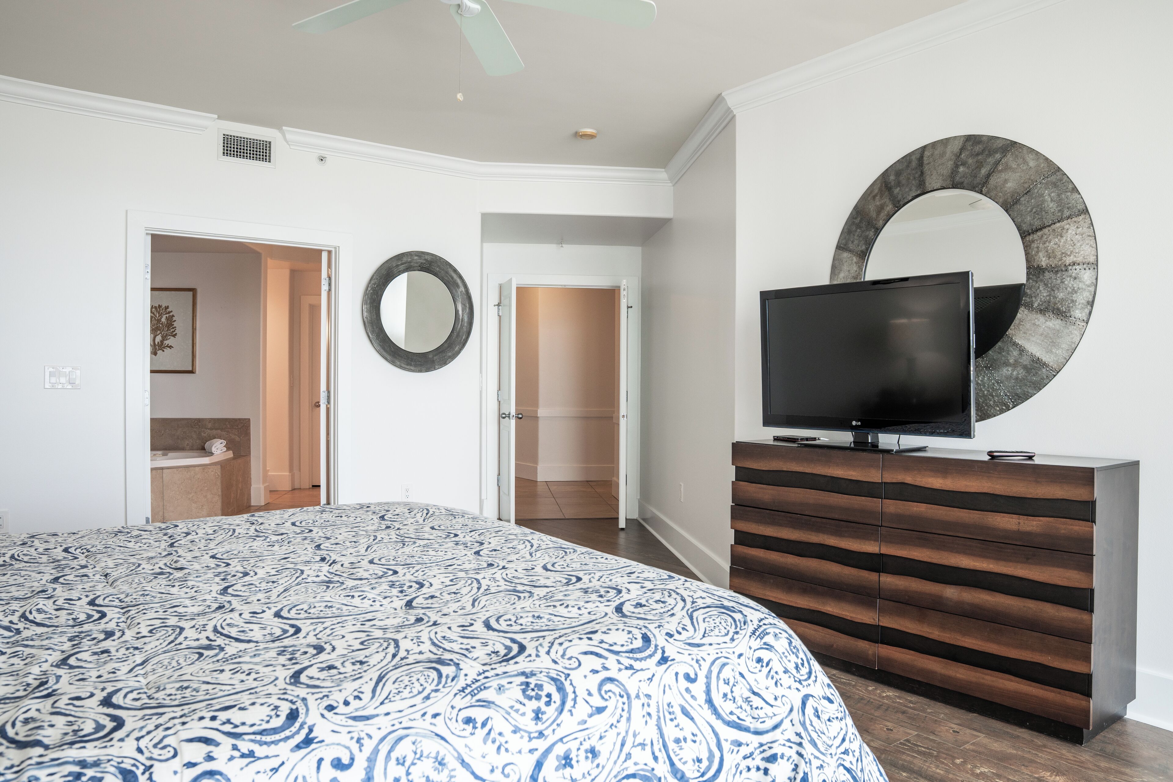 A bedroom with a patterned bedspread, a TV on a wooden dresser, a circular mirror, and a door leading to a bathroom.