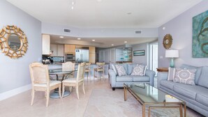A modern living room with a grey sofa, a glass coffee table, and a dining area with a glass table and wooden chairs. - Charming Beachy Condo Overlooking Perdido Bay and Gulf of Mexico~ Complimentary Boat Slip! (Orange Beach)
