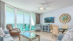 A spacious living area with a glass coffee table, comfortable seating, a ceiling fan, and a large window offering a view of the ocean. - Charming Beachy Condo Overlooking Perdido Bay and Gulf of Mexico~ Complimentary Boat Slip! (Orange Beach)