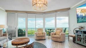 A spacious living room with a glass coffee table, two armchairs, and a view of the ocean through large sliding doors.
