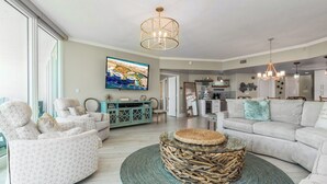 A spacious living room with a large TV, comfortable seating, and a kitchen in the background.
