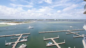 A marina with boats docked at piers, a sandy beach, and a distant cityscape under a partly cloudy sky. - Upscale Beach Condo Overlooking Robinson Island and Docks (Orange Beach)
