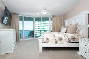 Relax in this serene bedroom with a large bed, dresser, ceiling fan, and TV, plus a sliding door to a balcony with a city view.