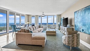 A spacious living room with a large sofa, ottoman, and armchair, a flat-screen TV, and a large abstract painting.