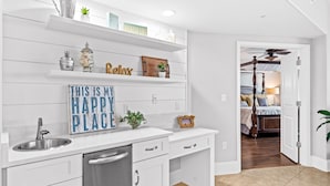 Una cucina moderna con mobili bianchi, lavello in acciaio inossidabile e lavastoviglie integrata. Gli scaffali sono decorati con diversi oggetti, incluso un cartello con la scritta "THIS IS MY HAPPY PLACE".
