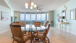 A spacious dining area with a round glass table surrounded by wicker chairs, large windows offering a view, and a ceiling fan. - Spacious Three Bedroom Condo with Wraparound View of Gulf! Complimentary Boat Slip! (Orange Beach)