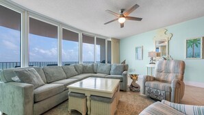A spacious living area with a large sectional sofa, a small round table, and a wicker chair. - Spacious Three Bedroom Condo with Wraparound View of Gulf! Complimentary Boat Slip! (Orange Beach)