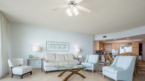Una sala de estar moderna con una mesa de centro de vidrio, un sofá y dos sillones. Se ve un área de cocina al fondo.
