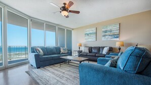 A spacious living room with a large window overlooking the beach, a ceiling fan, and two paintings on the wall. - Exquisite Three Bedroom Condo with Wraparound Balcony and Gulf Views! Complimentary Boat Slip! (Orange Beach)