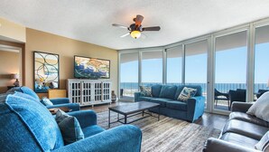 A spacious living room with a large sofa, a coffee table, and a television on a cabinet. The room has large windows offering a view of the ocean. - Exquisite Three Bedroom Condo with Wraparound Balcony and Gulf Views! Complimentary Boat Slip! (Orange Beach)
