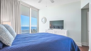 Ein Schlafzimmer mit einem großen Bett, einem Fernseher auf einer Kommode, einer Uhr an der Wand und Meerblick durch eine Schiebetür.
