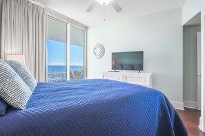 Un dormitorio con una cama grande, un televisor sobre un aparador, un reloj en la pared y vista al océano a través de una puerta corrediza.