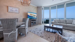 A living room with wicker furniture, a flat-screen TV, a coffee table, and a large window with a city view. - Relaxing Two Bedroom Penthouse with Gulf and Bay Views! Complimentary Boat Slip! (Orange Beach)