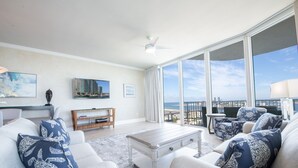 Un salón espacioso con un ventanal que ofrece vistas a la playa, un ventilador de techo y una chimenea moderna.
