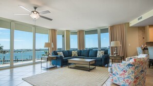 A spacious living room with a large window overlooking the sea, a ceiling fan, and a patterned armchair. - Spacious Unit with Wraparound Views of Bay and Quiet Courtyard! Complimentary Boat Slip! (Orange Beach)