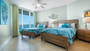 A bedroom with two wooden beds, a ceiling fan, a nightstand with a lamp, and a view of the sea through large windows. - Spacious Unit with Wraparound Views of Bay and Quiet Courtyard! Complimentary Boat Slip! (Orange Beach)