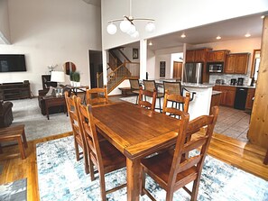 Uma sala de estar espaçosa com uma mesa e cadeiras de madeira, uma escada que leva a outro andar, e uma área de cozinha com armários de madeira e eletrodomésticos modernos.